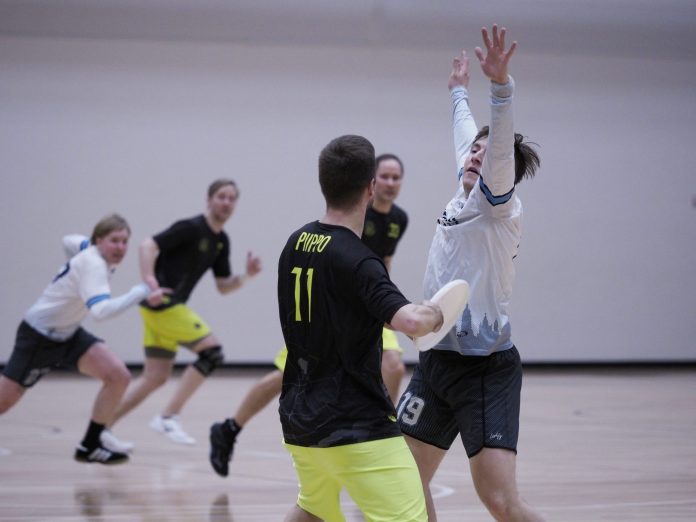 Pelaajalla on kiekko kädessä ja toinen pelaaja on kädet ylhäällä estämässä heittoa. Taustalla juoksee kaksi muuta pelaajaa.